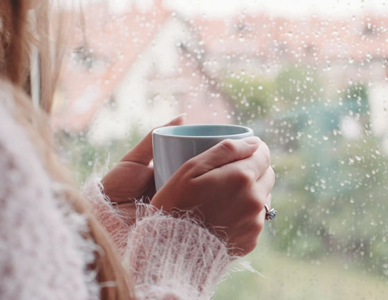 Mujer sosteniendo una taza con ambas manos mientras mira por la ventana en un día lluvioso