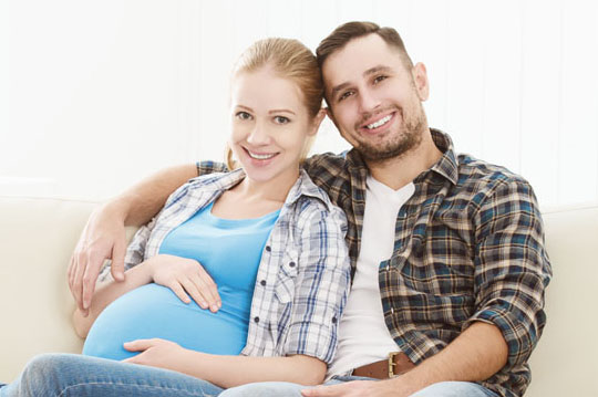 Pareja sentada en el sofá, la mujer embarazada y el hombre abrazándola con un brazo